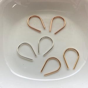 May include: Five U-shaped wire earrings arranged in a cluster. Two earrings are rose gold, two are gold, and one is silver. Each earring features a smooth, curved metal wire design with open ends, creating a simple, minimalist, and uniform shape.