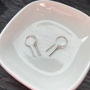 May include: A pair of silver-toned earrings, each featuring a circular hoop component with a short, straight bar pendant attached by a small jump ring. The earrings have a polished, metallic finish and a simple, clean design structure.
