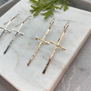 May include: A pair of gold cross earrings with a hammered texture. The earrings are long and dangle from the earlobe.
