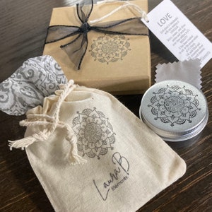 May include: A small, white, fabric pouch with a black mandala design and the text "Laura B Elements" printed on it. The pouch is sitting on a wooden surface next to a small, silver, round container with a black mandala design on the lid. The container is also sitting on a wooden surface. A small, brown box with a black ribbon and a black mandala design is in the background.
