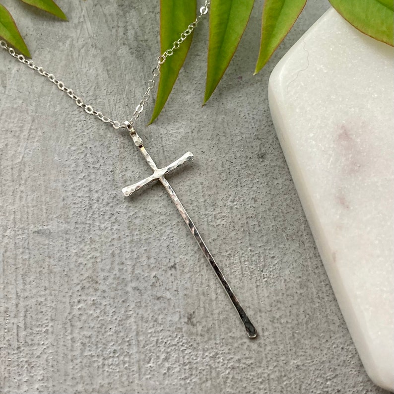 Pu&ograve; includere: Una collana con un ciondolo a forma di croce martellata in argento su una catena in argento. La croce &egrave; lunga e sottile con un'estremit&agrave; leggermente arrotondata.