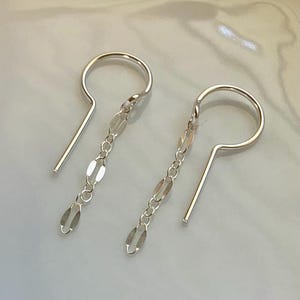 May include: A pair of silver earrings. Each earring features a curved top with a straight post and a chain of small, elongated oval links that dangle below. The earrings are displayed against a light-colored background.