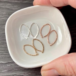 May include: Three pairs of marquise-shaped hoop earrings in silver, rose gold, and gold tones. Each earring features a delicate, hammered metal texture and a curved, open-loop design. The earrings are arranged inside a small, white ceramic dish.