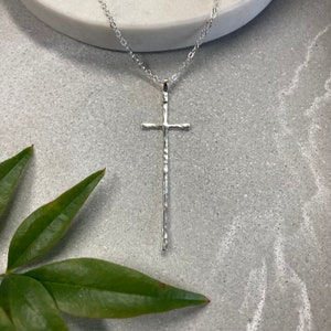 Pu&ograve; includere: Una collana con ciondolo a forma di croce in argento martellato. La croce &egrave; lunga e sottile con una superficie testurizzata.