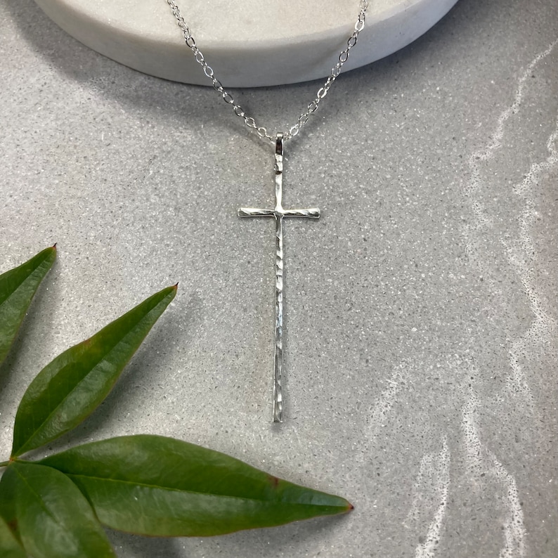 Pu&ograve; includere: Una collana con ciondolo a forma di croce in argento martellato. La croce &egrave; lunga e sottile con una superficie testurizzata.