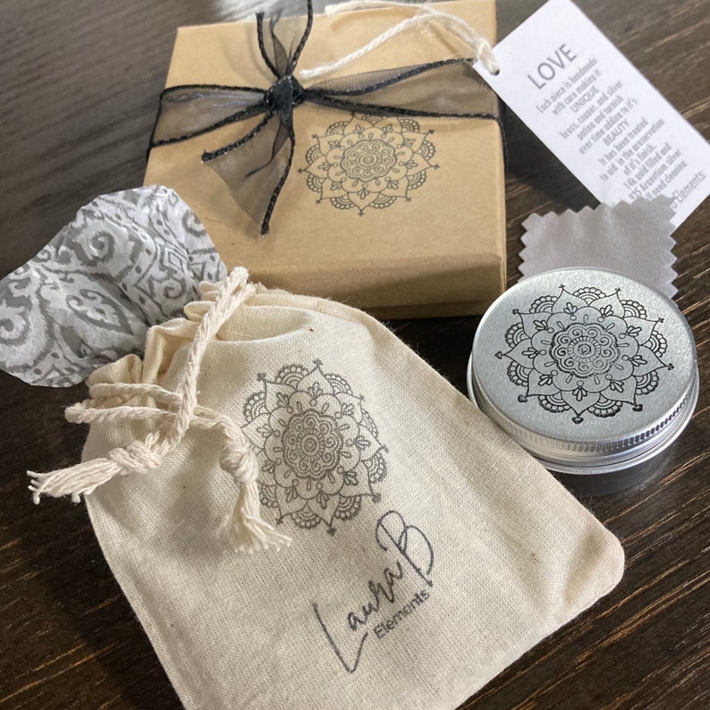 Pu&ograve; includere: Una piccola borsa di stoffa bianca con un disegno di mandala nero e il testo "Laura B Elements" stampato su di essa. La borsa &egrave; appoggiata su una superficie di legno accanto a un piccolo contenitore argentato con un disegno di mandala nero sul coperchio. Il contenitore &egrave; anche appoggiato su una superficie di legno. C'&egrave; una piccola scatola marrone con un nastro nero e un disegno di mandala nero sul coperchio. La scatola &egrave; appoggiata su una superficie di legno. C'&egrave; un piccolo panno bianco con un motivo grigio sulla superficie. Il panno &egrave; appoggiato su una superficie di legno. C'&egrave; un piccolo biglietto bianco con il testo "LOVE" stampato su di esso. Il biglietto &egrave; appoggiato su una superficie di legno.