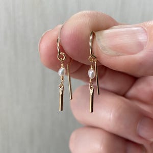 May include: Delicate gold-colored drop earrings. Each earring features a small, white pearl and a slender, gold-colored bar. The earrings are held up by a curved hook. The design is simple and elegant, suitable for everyday wear or special occasions.