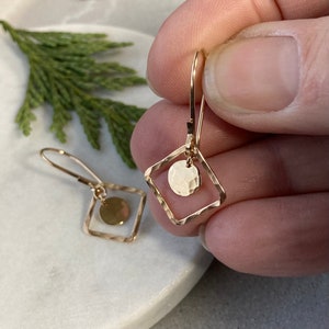 May include: Gold-colored earrings with a hammered finish. Each earring features a square frame with a small, hammered disc suspended inside. The earrings are held up by a lever back closure.