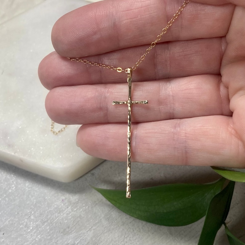 Pu&ograve; includere: Una collana con catena d'oro con un ciondolo a forma di croce lunga e martellata.