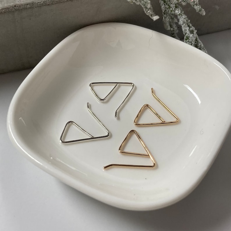 May include: Four triangle-shaped earring hooks, two gold and two silver, displayed on a white ceramic dish.