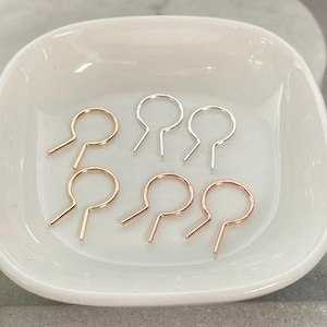 May include: A collection of six metal stitch markers in a white ceramic dish. The stitch markers are in three different colors: gold, silver, and rose gold. Each marker has a circular top with two straight prongs extending from the bottom.
