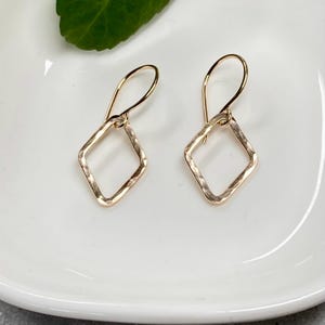 May include: A pair of gold-colored earrings. Each earring features a hammered, diamond-shaped pendant suspended from a curved ear wire. The earrings are displayed on a white surface, with a green leaf visible in the background.