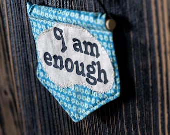 May include: A blue and white fabric pennant with the words "I am enough" printed in black on a white background. The pennant is hanging on a dark wood surface.
