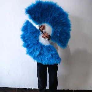 May include: Two large blue feather fans held in front of a person's face. The fans are made of soft, fluffy feathers and are held open in a semi-circle shape.