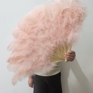May include: A large, open fan with light pink feathers. The feathers are arranged in layers, creating a full, fluffy appearance. The handle is made of light-coloured wood. The fan is held in front of a person.
