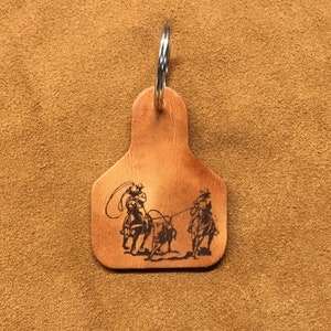 May include: A brown leather keychain shaped like a cattle ear tag with a metal ring. The keychain features an engraved image of two cowboys on horseback roping a calf.