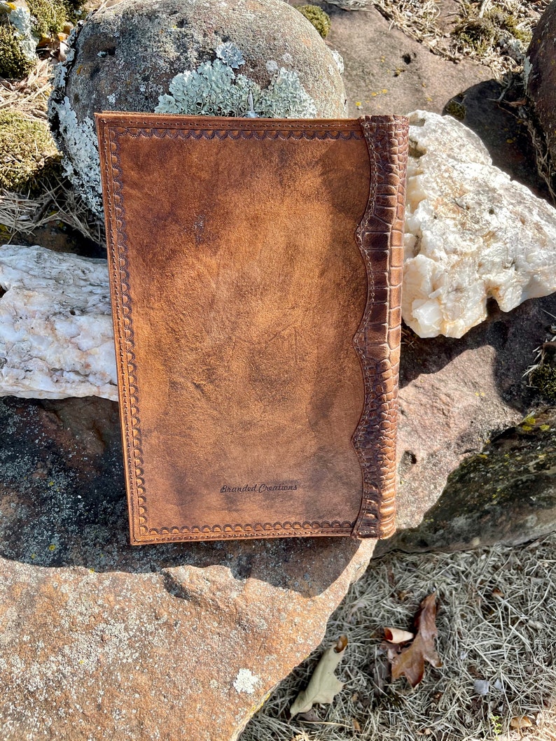 May include: A brown leather journal with a decorative embossed border and a spine with a crocodile pattern. The journal has the text "Branded Creations" embossed on the front cover.