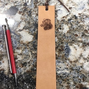 Op de afbeelding: Een bruine leren bladwijzer met een gegraveerd silhouet van een zwarte en bruine hond. De bladwijzer heeft een bruine leren kwast.