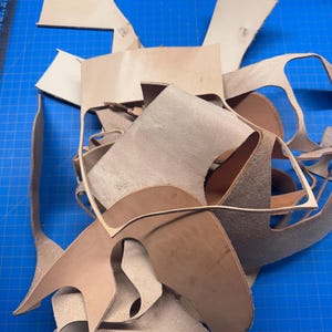 May include: A collection of irregularly shaped leather pieces in various shades of tan and brown. The pieces are scattered on a blue cutting mat, suggesting a crafting or leatherworking project. Some pieces have curved edges.