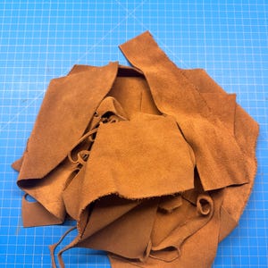 Può includere: Un mucchio di pezzi di pelle scamosciata marrone, alcuni con bordi tagliati e lacci, sono disposti su un tappetino da taglio blu a griglia. La pelle ha un aspetto morbido e strutturato, adatta per l'artigianato o l'abbigliamento.