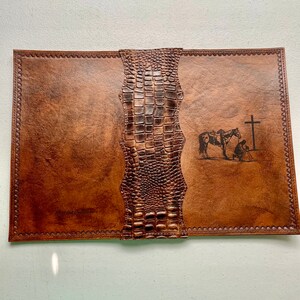 May include: A brown leather journal with a crocodile embossed spine. The cover features a design of a person on horseback kneeling before a cross. The text "Blessed Beginnings" is embossed on the front cover.