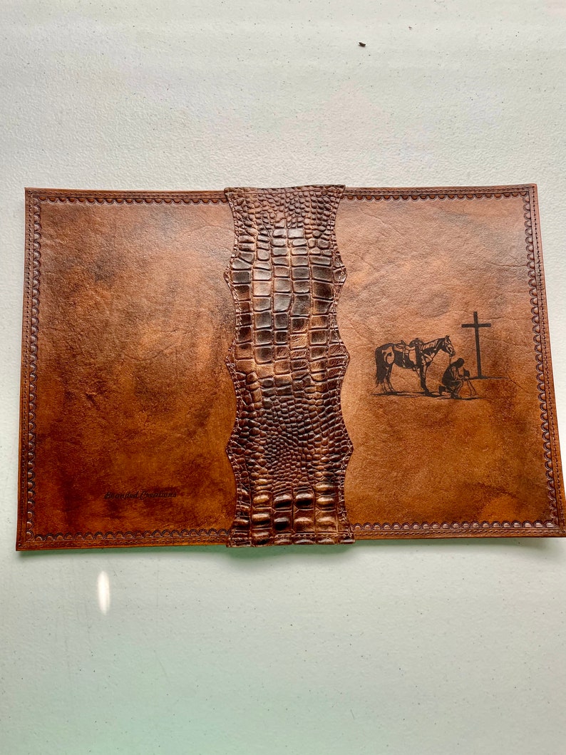 May include: A brown leather journal with a crocodile embossed spine. The cover features a design of a person on horseback kneeling before a cross. The text "Blessed Beginnings" is embossed on the front cover.