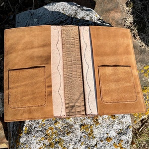 May include: A brown leather journal with two pockets on the front and back covers. The journal has a decorative stitched design on the spine.