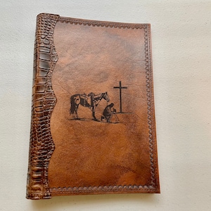 May include: A brown leather journal with a decorative embossed border and a cowboy on horseback praying at a cross design. The journal has a crocodile-embossed spine.