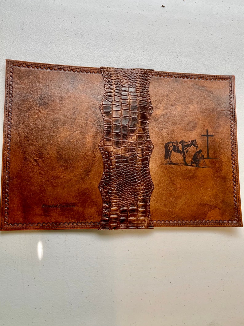 May include: A brown leather journal with a crocodile embossed cover. The journal has a cross and a horse with a rider engraved on the front cover. The text "Blessed Treasures" is embossed on the front cover.