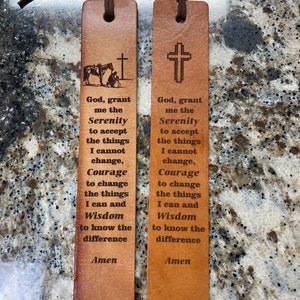 May include: Two leather bookmarks with brown suede ties. One bookmark features a cross and the other depicts a cowboy kneeling before a cross with a horse. Both have the text "God, grant me the Serenity..." and "Amen".