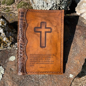 May include: A brown leather-bound journal with a cross embossed on the front cover. The journal has decorative stitching around the edges and a quote from Joshua 1:9 is inscribed below the cross.