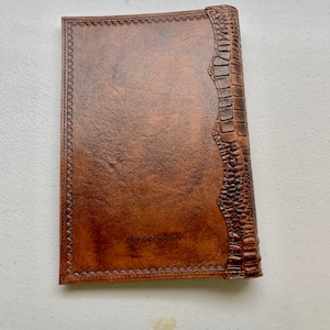 May include: A brown leather-bound journal with a decorative embossed border and a crocodile-embossed spine. The journal has the text "Branded Creations" embossed on the front cover.