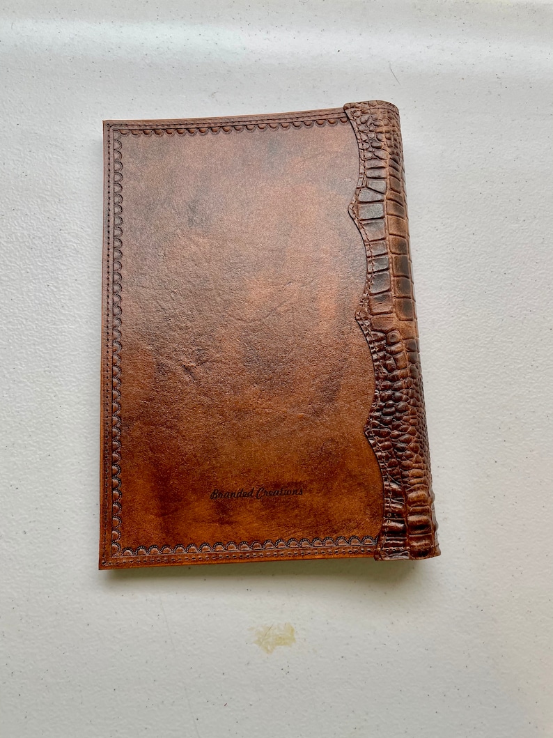 May include: A brown leather-bound journal with a decorative embossed border and a crocodile-embossed spine. The journal has the text "Branded Creations" embossed on the front cover.