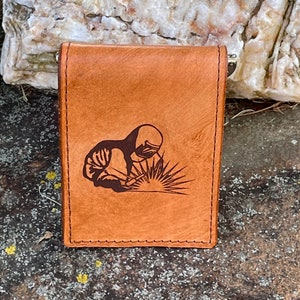 May include: A brown leather wallet with a stitched border. The front features a dark brown illustration of a welder at work. The wallet is rectangular and appears to be closed.