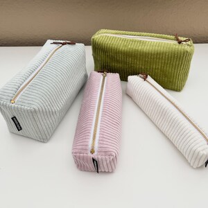 Può includere: Quattro astucci rettangolari in velluto a coste nei colori azzurro, verde oliva, rosa antico e bianco sporco. Ogni astuccio presenta una chiusura a zip in metallo dorato con una piccola linguetta in pelle marrone e un'etichetta nera sul lato.