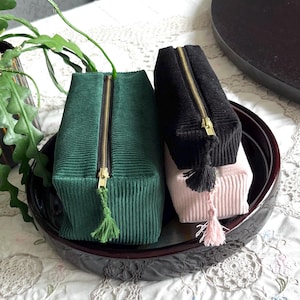 Può includere: Tre pochette con cerniera in velluto a coste verde, nero e rosa, ciascuna con cerniera e nappa color oro. Le pochette sono rettangolari e si trovano all'interno di un vassoio rotondo e scuro. La pochette verde è in primo piano.