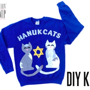 Può includere: Una felpa blu con la scritta "HANUKCATS" in lettere bianche. Due gatti di feltro, uno grigio e uno bianco, sono sulla felpa con una stella di David gialla tra loro. La felpa è un kit fai-da-te.