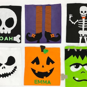 May include: Six Halloween-themed fabric bags with different characters. The bags are black, white, purple, orange, and green. The characters include a skull, witch's legs, a skeleton, a pumpkin, and Frankenstein. The bags have the names "Noah" and "Emma" written on them.