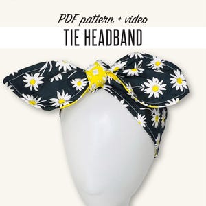 May include: A tie headband with a black background and white daisy pattern, accented with yellow trim. The headband is displayed on a white mannequin head. Text on the image reads "PDF pattern + video TIE HEADBAND". The Fashion Camp logo is in the upper left corner.