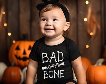 May include: A baby wearing a black bodysuit with the text "Bad to the Bone" in white. The baby is wearing a black bow headband and is sitting in front of a pumpkin.