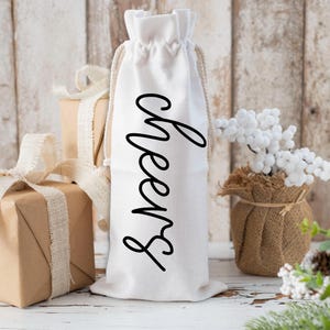 Könnte beinhalten: Eine weiße Weinflaschentasche mit dem Wort "Cheers" in schwarzer Schrift. Die Tasche hat einen Kordelzugverschluss und eine beige Schnur. Braune Geschenkverpackungen mit beigem Band befinden sich im Hintergrund, zusammen mit einem kleinen Jutesack mit weißen Beeren.