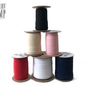 May include: Six spools of different colored elastic fabric are stacked on top of each other. The spools are labeled with the text "FASHION CAMP create. design. sew."