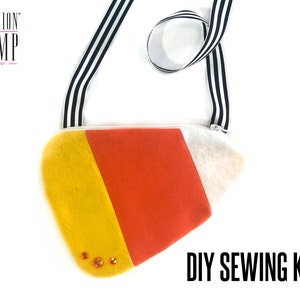 May include: DIY sewing kit to make a candy corn-shaped crossbody bag. The bag is made of felt in yellow, orange, and white. It has a black and white striped strap and a zipper closure. The bag is decorated with orange rhinestones.