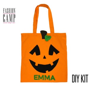 Peut inclure: Sac fourre-tout en tissu orange avec un visage de citrouille en feutre noir et une tige en feutre vert. Le sac porte l'inscription "FASHION CAMP" et "DIY KIT". Le nom "EMMA" est &eacute;crit en feutre vert sur le devant du sac.