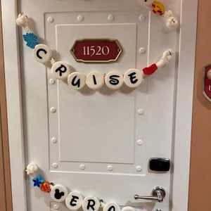May include: White door decorated with garlands spelling "In Our Cruise Era". The garlands feature decorative elements like Mickey Mouse silhouettes and colorful accents. The door has a room number plaque.