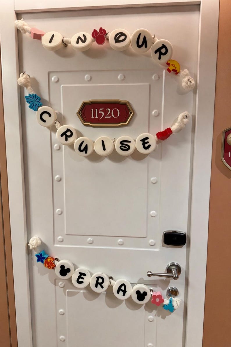May include: White door decorated with garlands spelling "In Our Cruise Era". The garlands feature decorative elements like Mickey Mouse silhouettes and colorful accents. The door has a room number plaque.