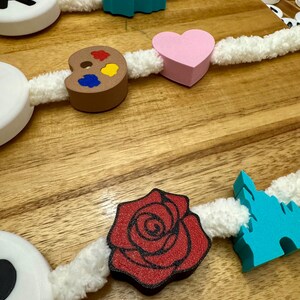 May include: A decorative garland featuring various wooden shapes, including a red rose, a pink heart, and a blue firework design. The shapes are strung together with a white, fluffy cord, creating a whimsical and colorful aesthetic. The garland is displayed on a wooden surface.