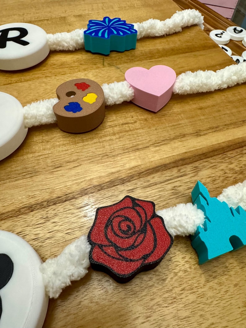 May include: A decorative garland featuring various wooden shapes, including a red rose, a pink heart, and a blue firework design. The shapes are strung together with a white, fluffy cord, creating a whimsical and colorful aesthetic. The garland is displayed on a wooden surface.