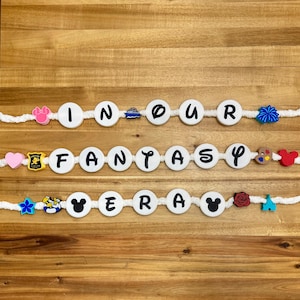 May include: Three rows of white beads spell out "IN OUR FANTASY ERA" with decorative charms. The charms include a pink bow, a ship, a blue flower, a heart, a castle, a Mickey Mouse head, and a rose.