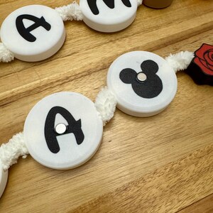 May include: A decorative garland featuring white circular beads with black letters and a Mickey Mouse silhouette. Additional elements include a wooden artist's palette and a red rose. The garland is strung on a white, fuzzy cord.